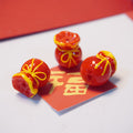 Three glossy red decorative beads shaped like wrapped red envelopes, with yellow trim and bow details, arranged on a red card over a white surface.