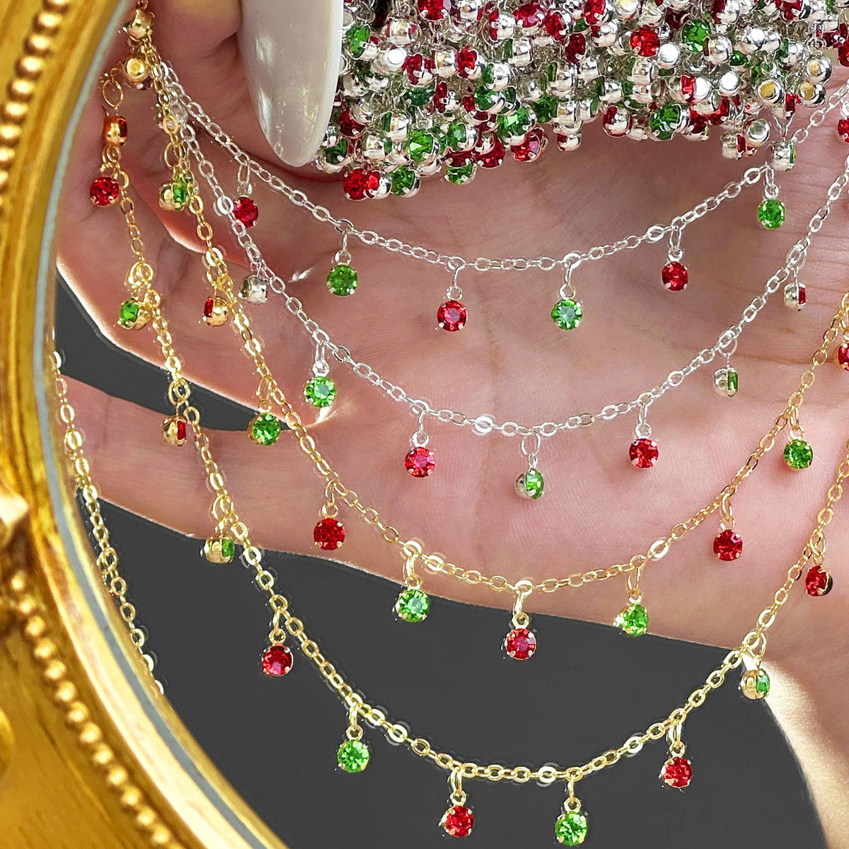 Three layered dangle chain necklaces in gold, silver, and white metals, embellished with red and green rhinestones, draped over a hand.