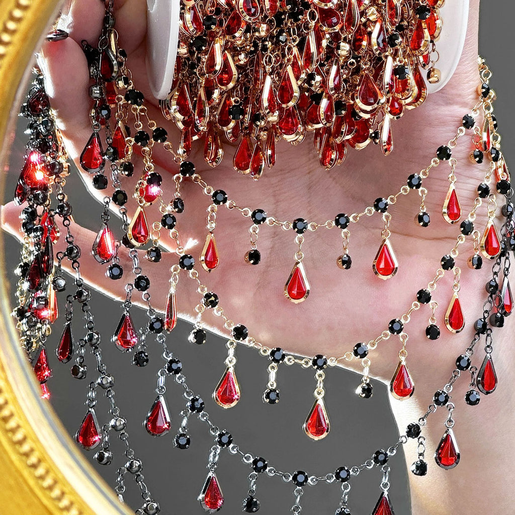 A close-up of a hand holding a large gold-framed mirror that reflects a dense, layered arrangement of red teardrop crystal drops, black faceted beads, and gold and silver chain drapes.