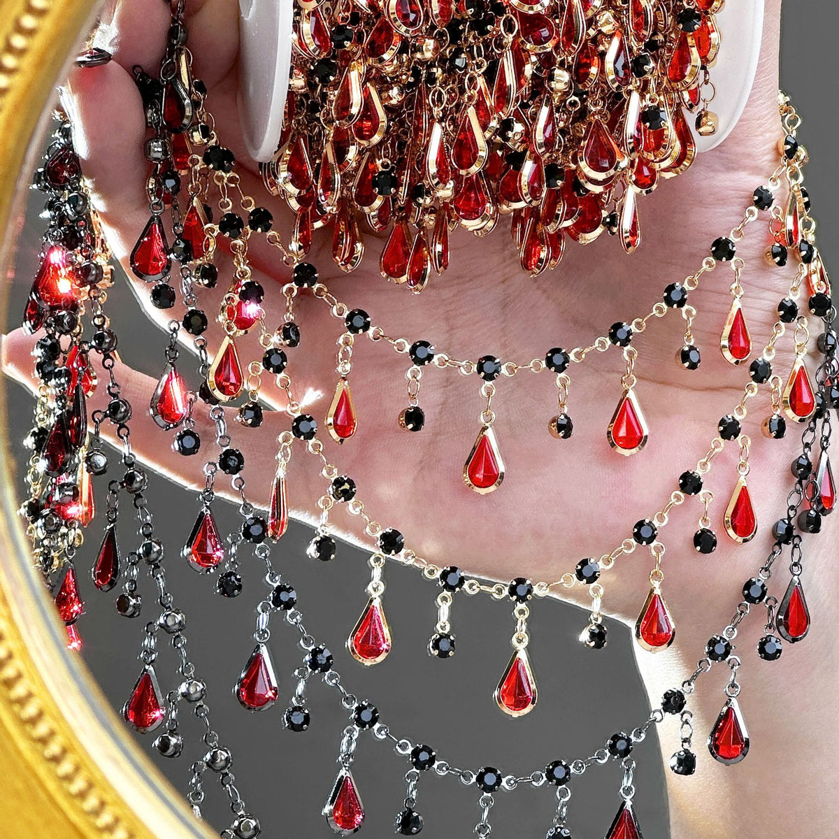 A close-up of a hand holding a large gold-framed mirror that reflects a dense, layered arrangement of red teardrop crystal drops, black faceted beads, and gold and silver chain drapes.