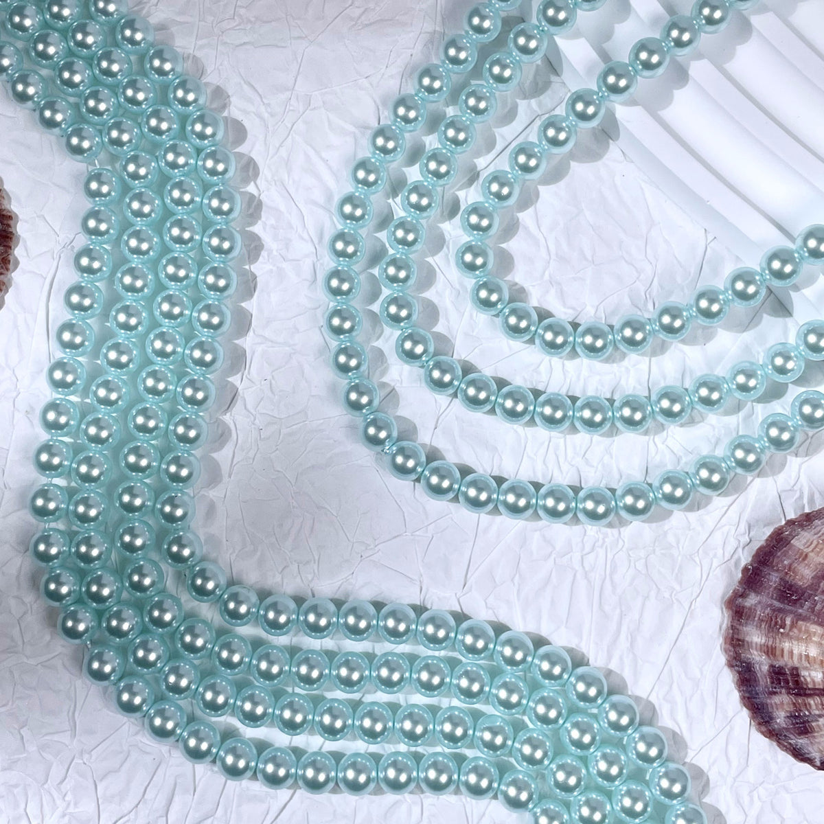 Light blue translucent mesh trim with small pearl-like beads, arranged in loops on a white textured surface with seashell props.
