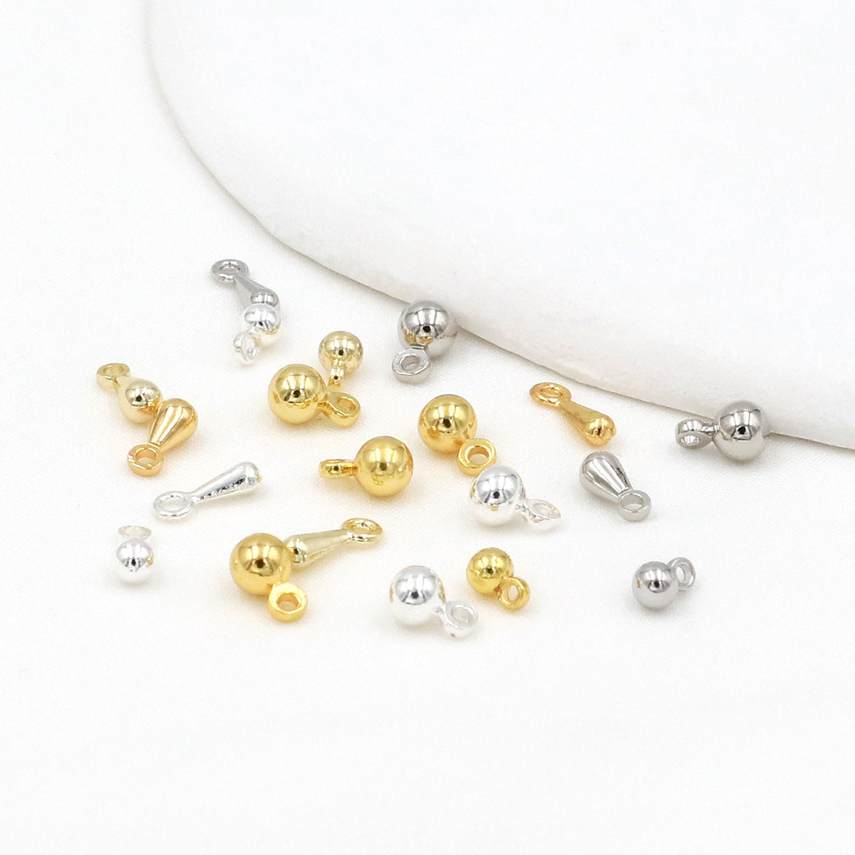 Gold and silver ball and teardrop charms scattered on a white surface with a curved white prop