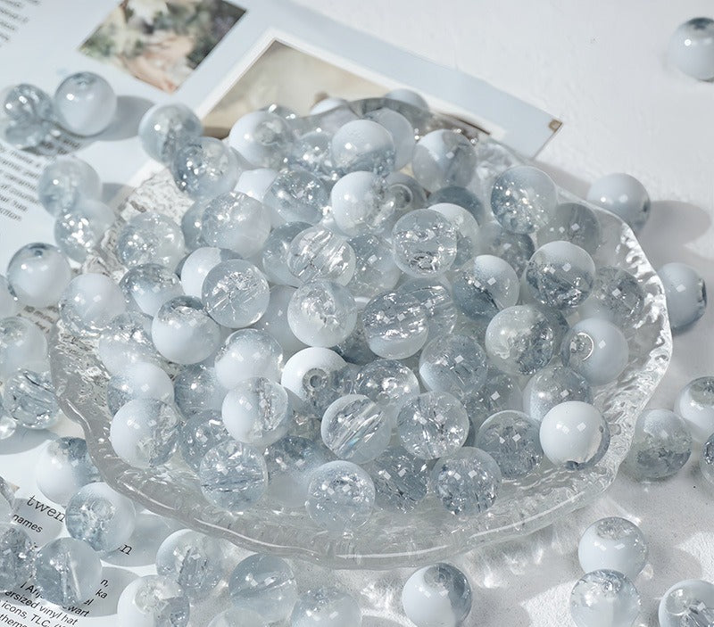 Clear and white ice-crackle glass beads piled in a clear glass dish on a white surface with scattered beads