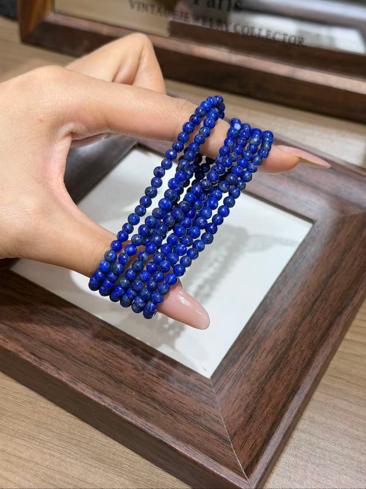 Hand with long manicured nails holding a multi-strand royal blue beaded bracelet over a dark wooden frame on a wooden table.