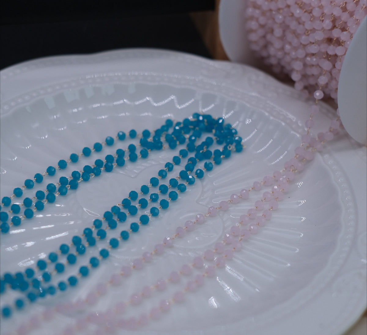 Two strands of faceted round beads, one in bright turquoise blue and one in light pink, laid out on a white textured ceramic plate.