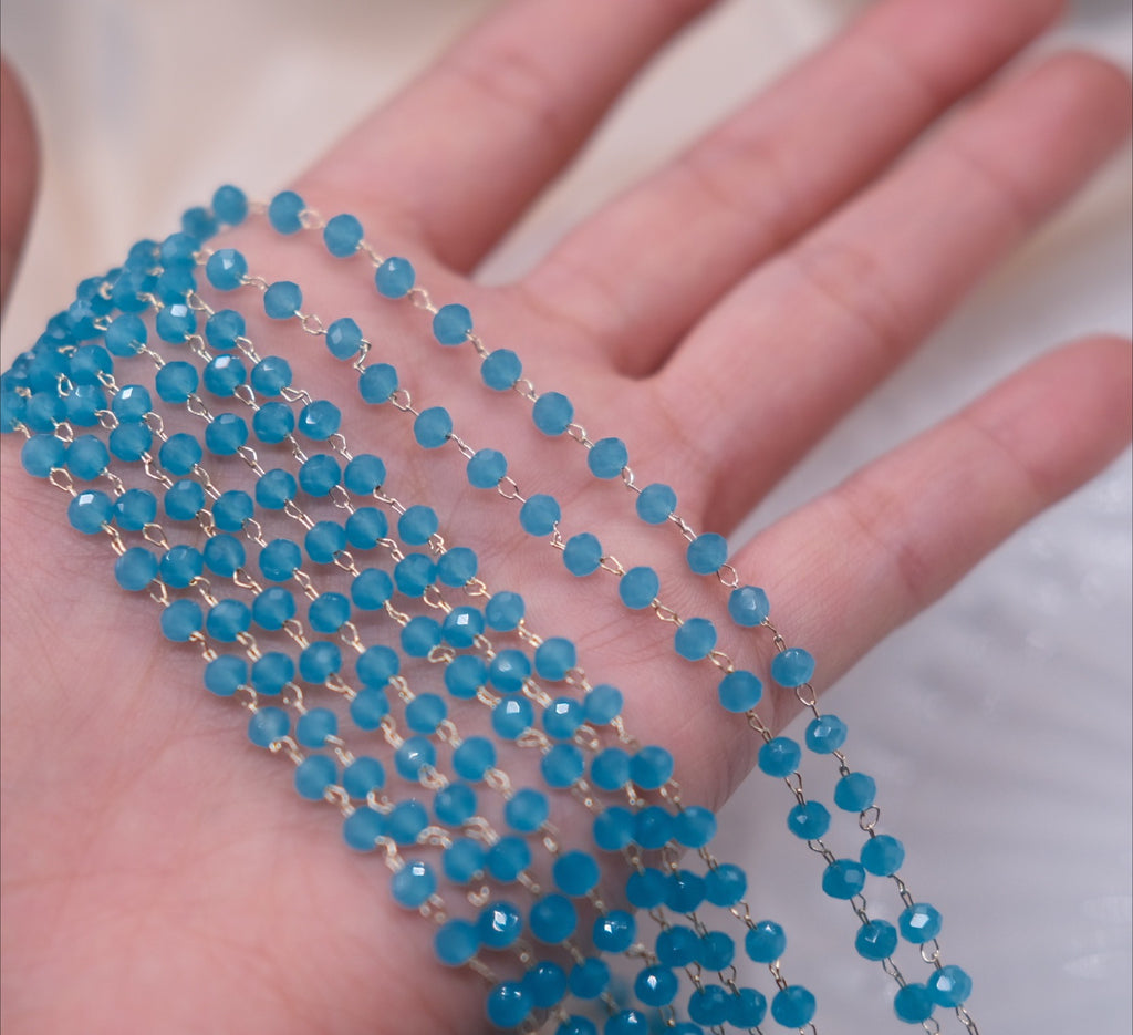 A close-up of a hand holding multiple strands of faceted blue beads connected by small gold-toned metal links, shown against a soft light background.