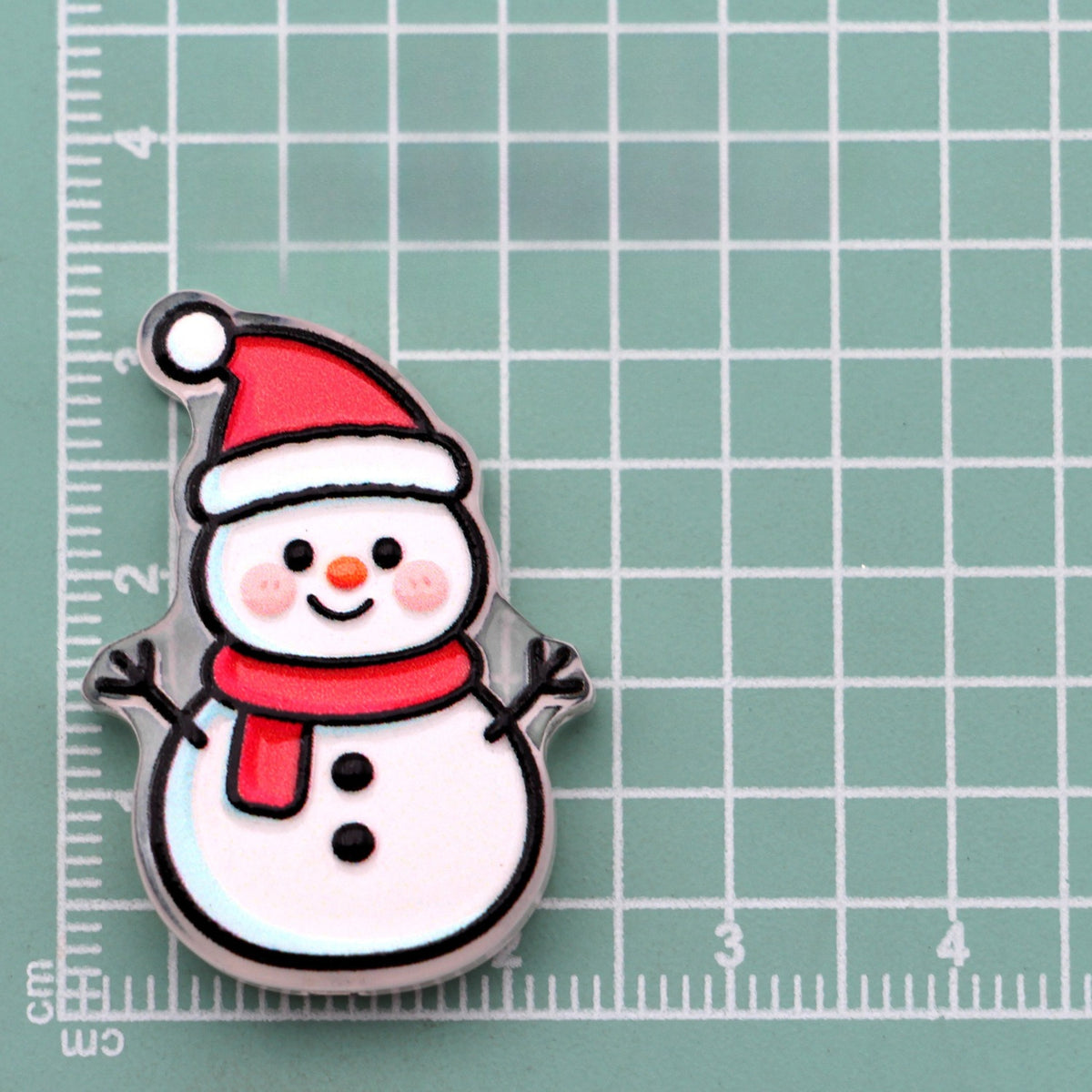 Snowman acrylic charm with red Santa hat and red scarf on green cutting mat