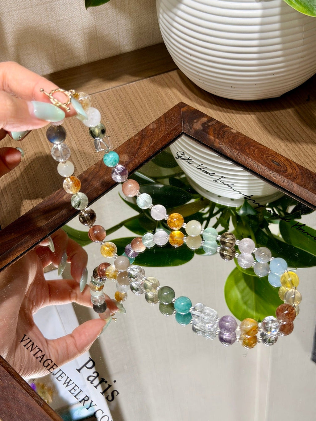 Hands holding multicolor faceted bead necklace reflected in mirror, white vase and green leaves behind