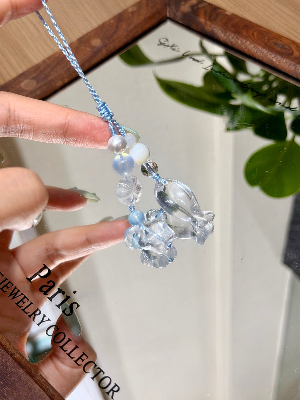 Hand holding light blue cord phone charm with clear beads and glass flower against mirror.