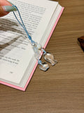 Hand holding blue cord phone charm with white beads and fish charms over open book.