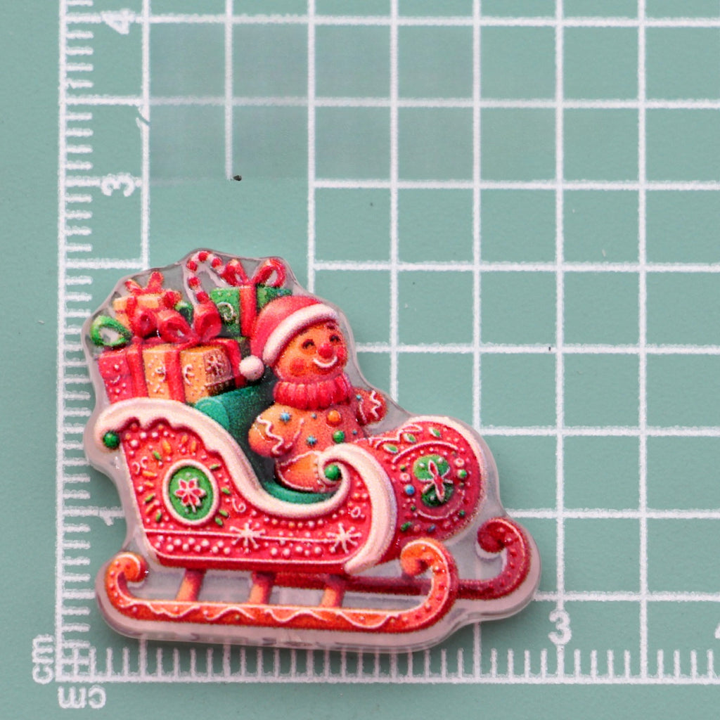 Gingerbread man in Santa hat riding red sleigh acrylic charm with presents on green cutting mat
