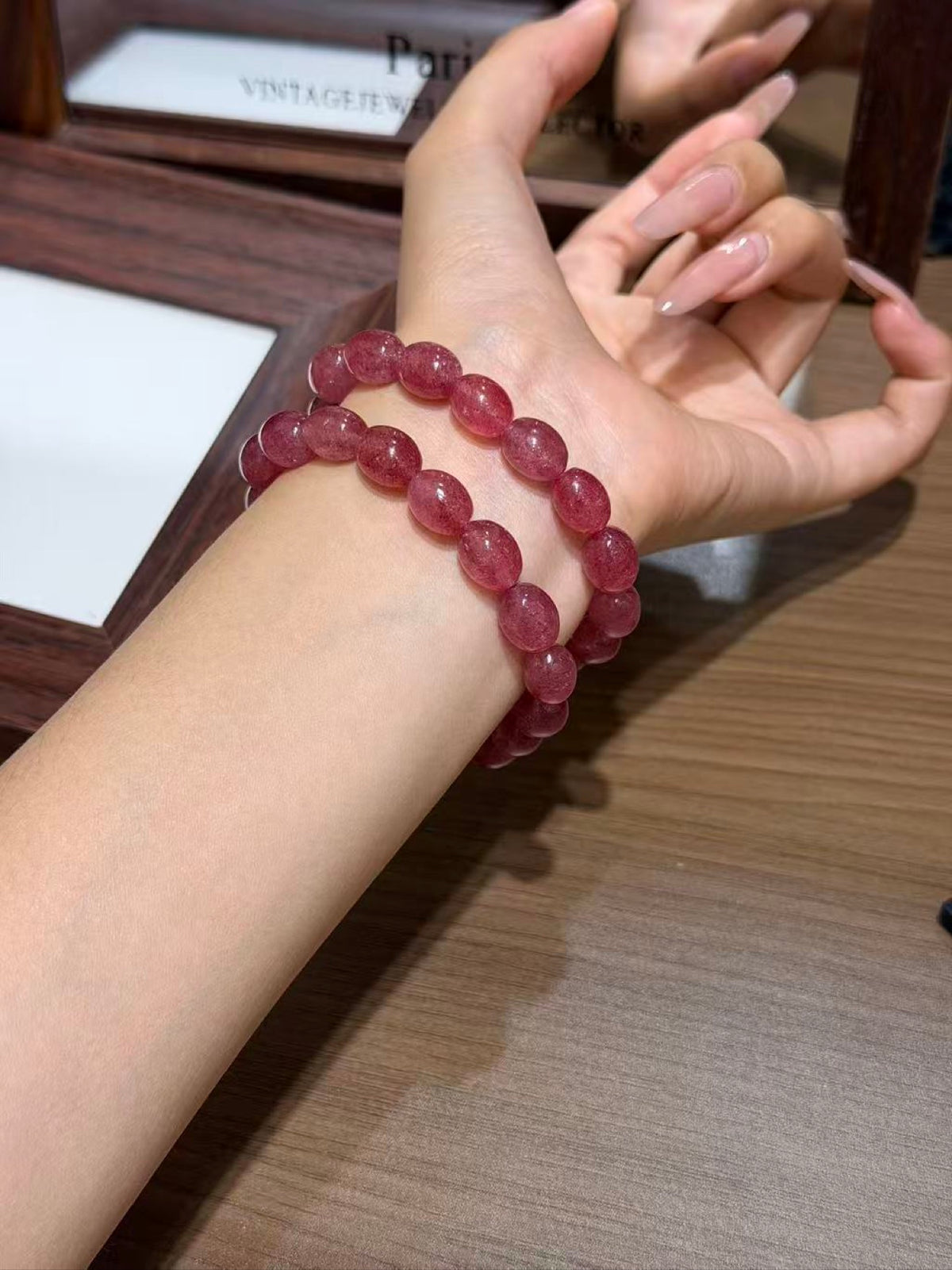 Strawberry quartz jelly bean bracelet of translucent red-pink egg beads worn on a wrist, hand with long manicured nails raised near the face, over a wooden tabletop