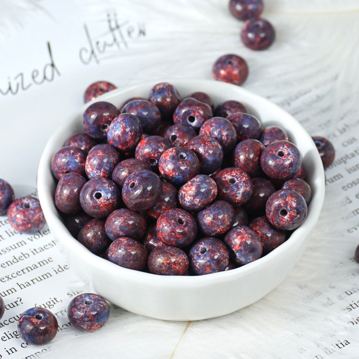 White porcelain bowl filled with round dark purple and red mottled glazed porcelain beads, loose beads on printed paper