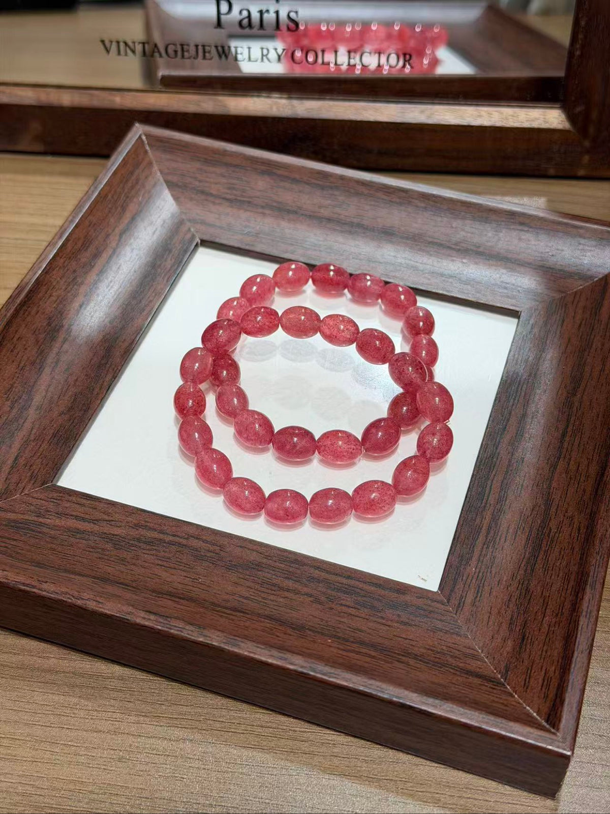 Strawberry quartz jelly bean bracelet of red-pink egg beads coiled on a white insert inside a dark brown wooden frame, with a blurred mirror behind showing the text 'Paris Vintage Jewelry Collector'