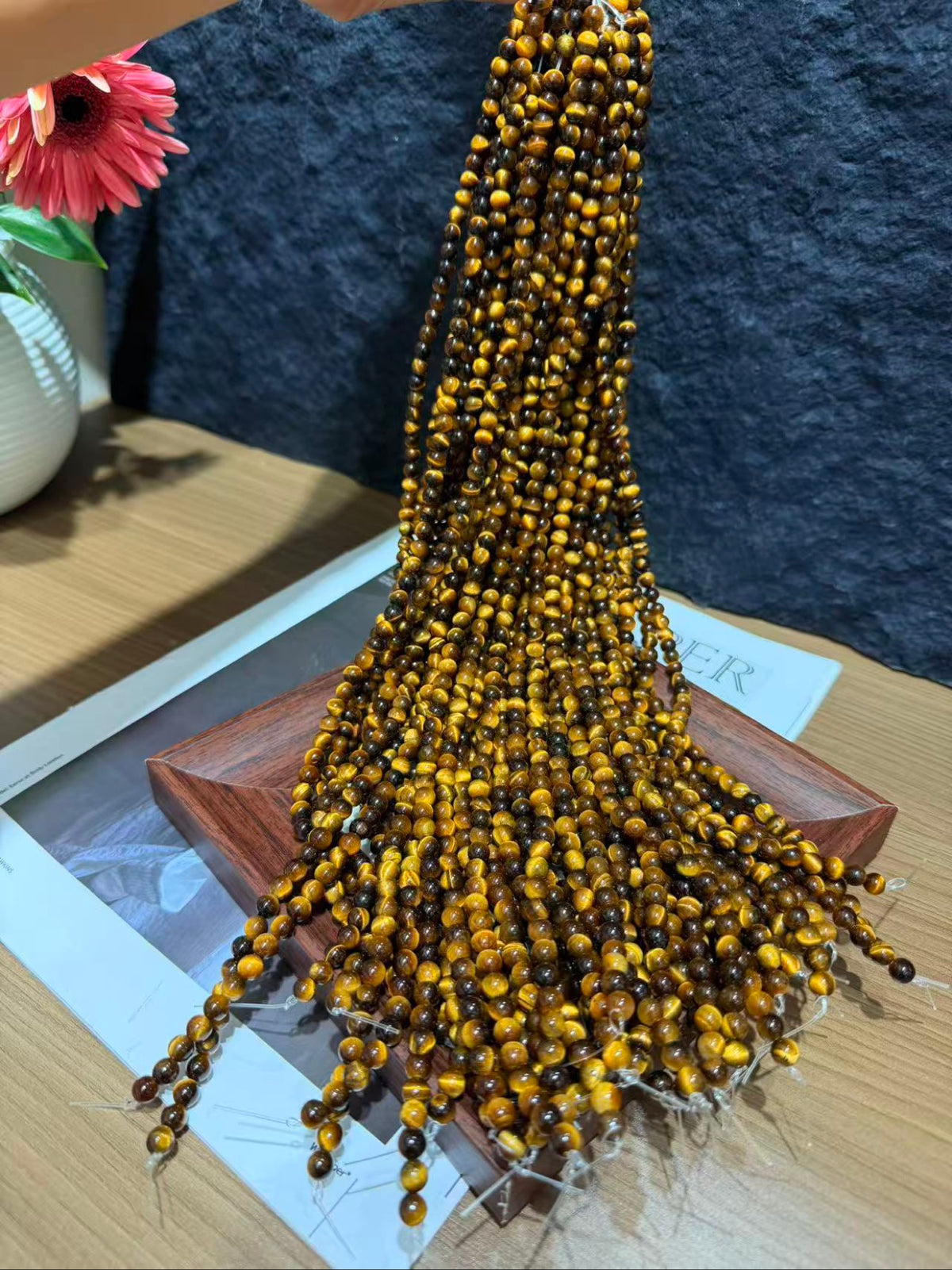 Bundle of long strands of yellow tiger's eye beads cascading over a display, with a pink flower and dark textured wall in the background