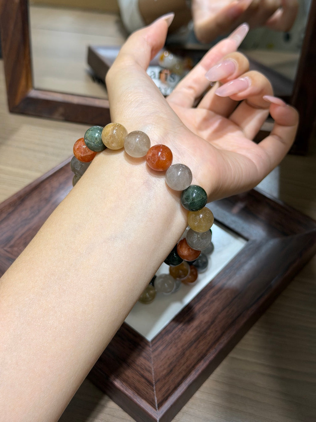 A person's wrist wearing the multicolor rabbit-hair quartz bead bracelet with round polished stones in green, orange, yellow, and translucent white, above a dark wooden tray