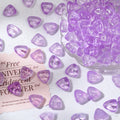 Purple translucent triangular glass beads scattered on a white surface with a clear glass bowl of beads