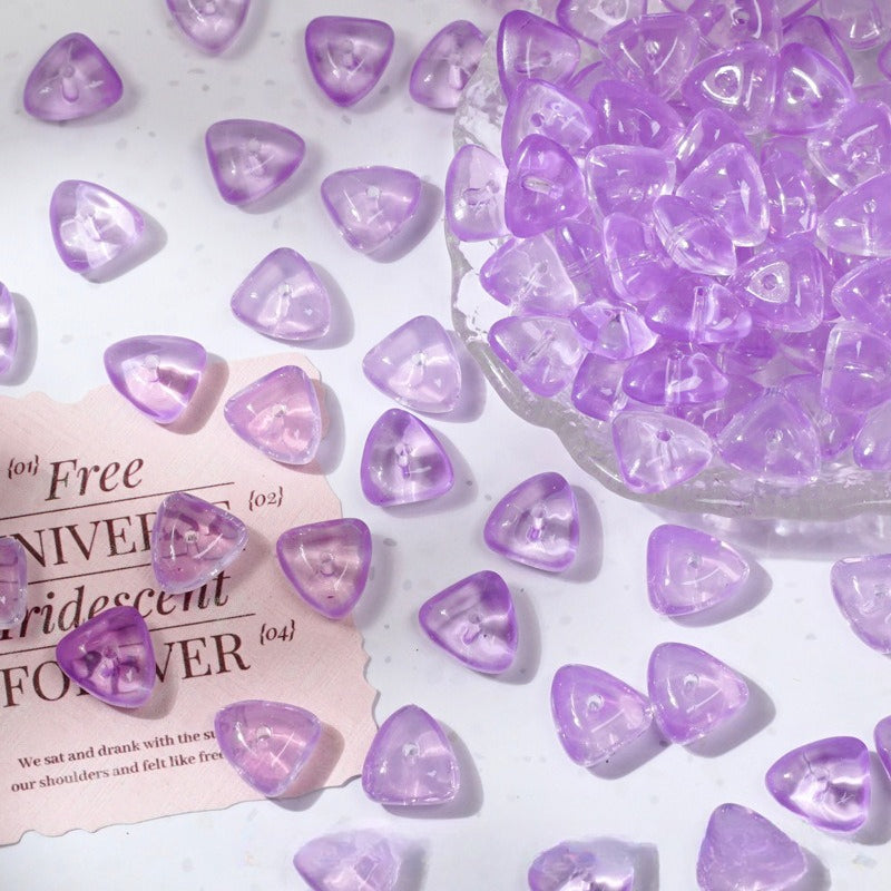 Purple translucent triangular glass beads scattered on a white surface with a clear glass bowl of beads