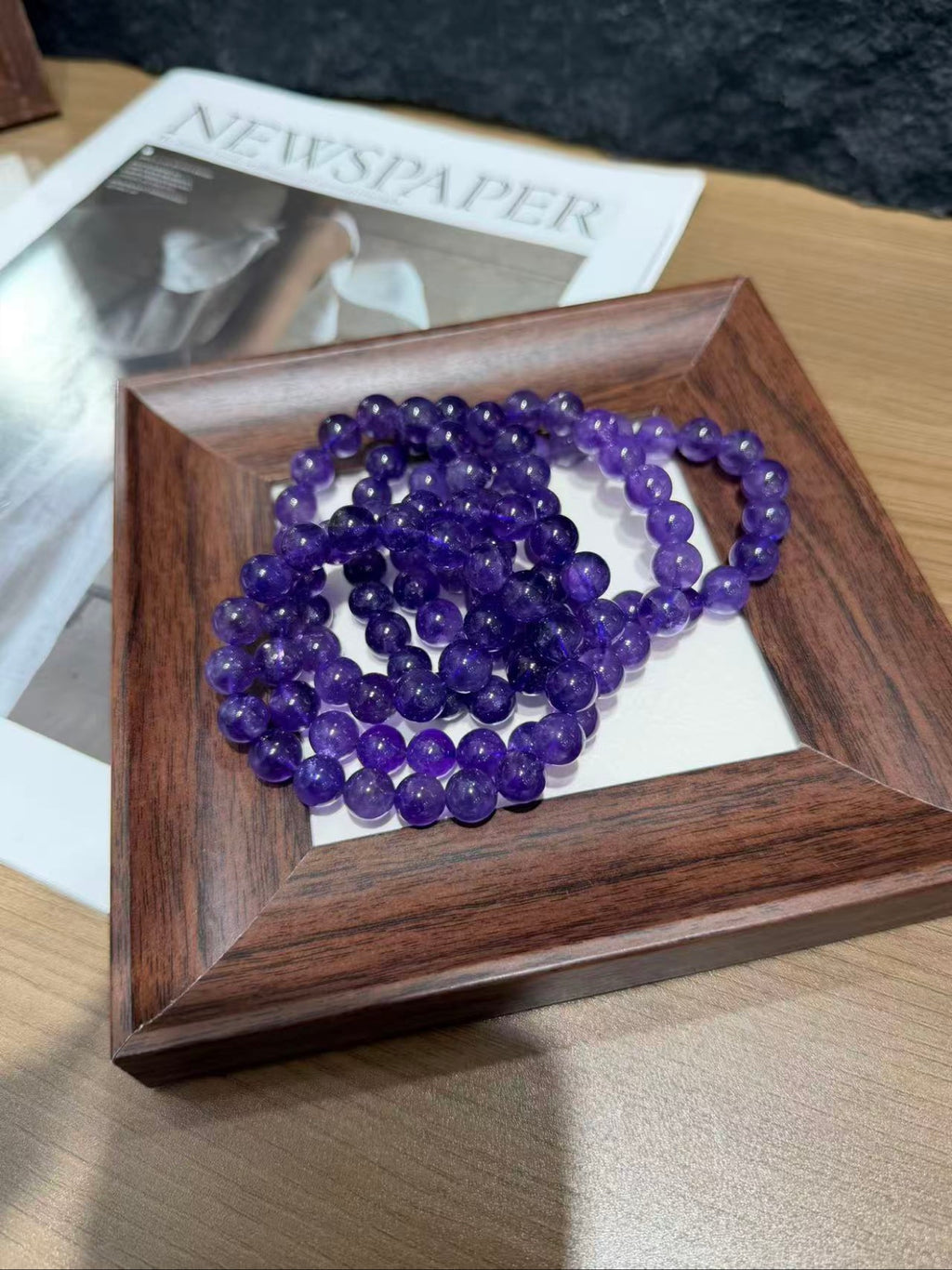 Coiled purple amethyst bead bracelets in a wooden display tray on a table with a newspaper underneath