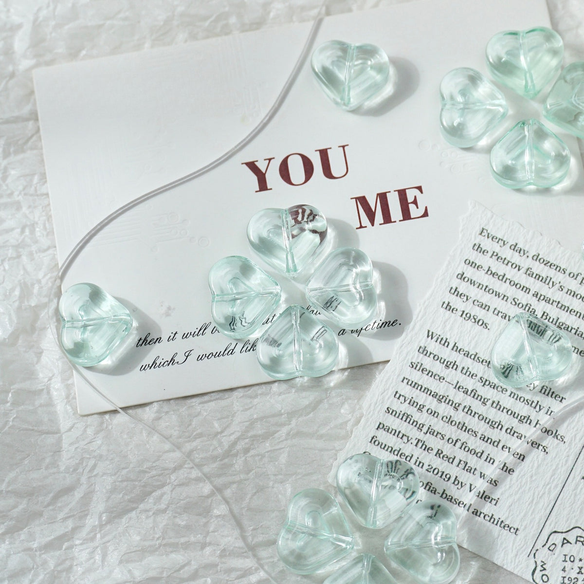 Clear glass heart-shaped and flower-shaped beads with a silver wire, arranged on a white textured fabric alongside printed cards.