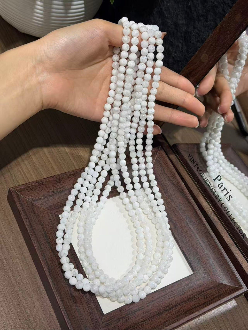 Hand holding multiple strands of white snowflake phantom quartz round beads over a dark wooden display