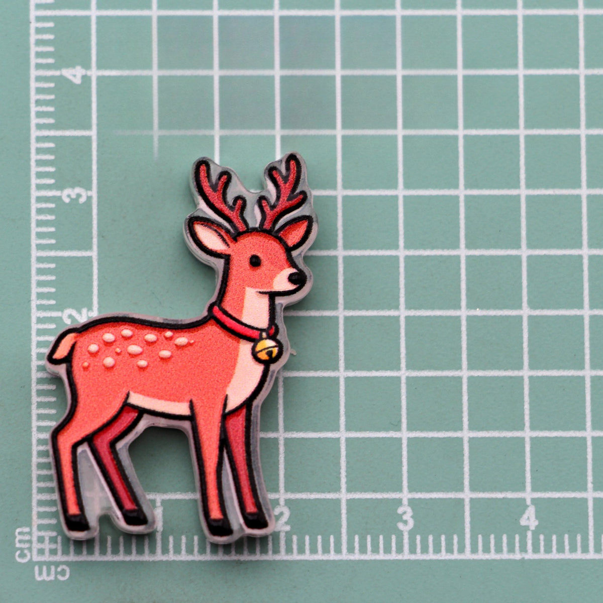 Reindeer acrylic charm with red antlers, red collar, and gold bell on green cutting mat