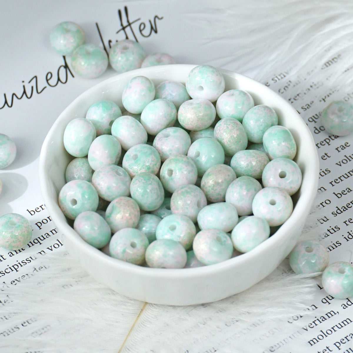 Round beads with mottled pastel mint green, white and pink glitter finish in a small white porcelain bowl on printed paper
