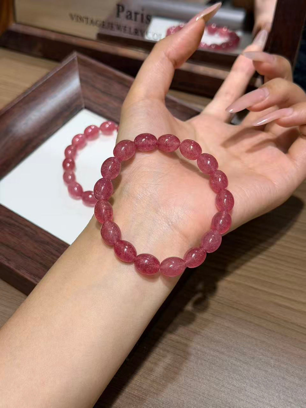 Pink strawberry quartz jelly bean bracelet worn on a wrist with the palm facing up, another bracelet resting on a white insert in a dark wooden tray behind, mirror reflection showing 'Paris Vintage Jewelry Collector'