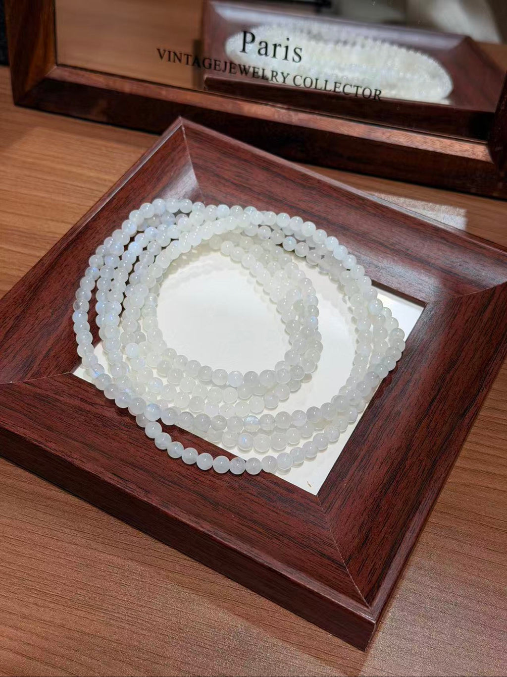 Translucent pale blue-white beaded bracelets coiled in a dark wooden display tray, with a matching wooden box labeled "Paris Vintage Jewelry Collector" in the background