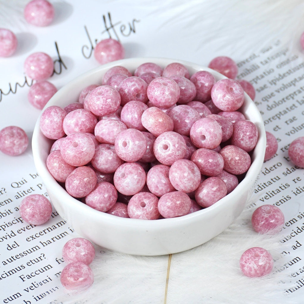 Pink glittery round beads with holes in a small white porcelain bowl, some scattered on printed paper