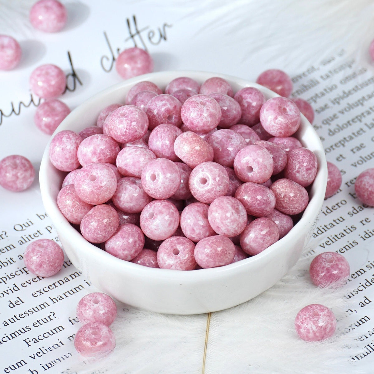 Pink glittery round beads with holes in a small white porcelain bowl, some scattered on printed paper
