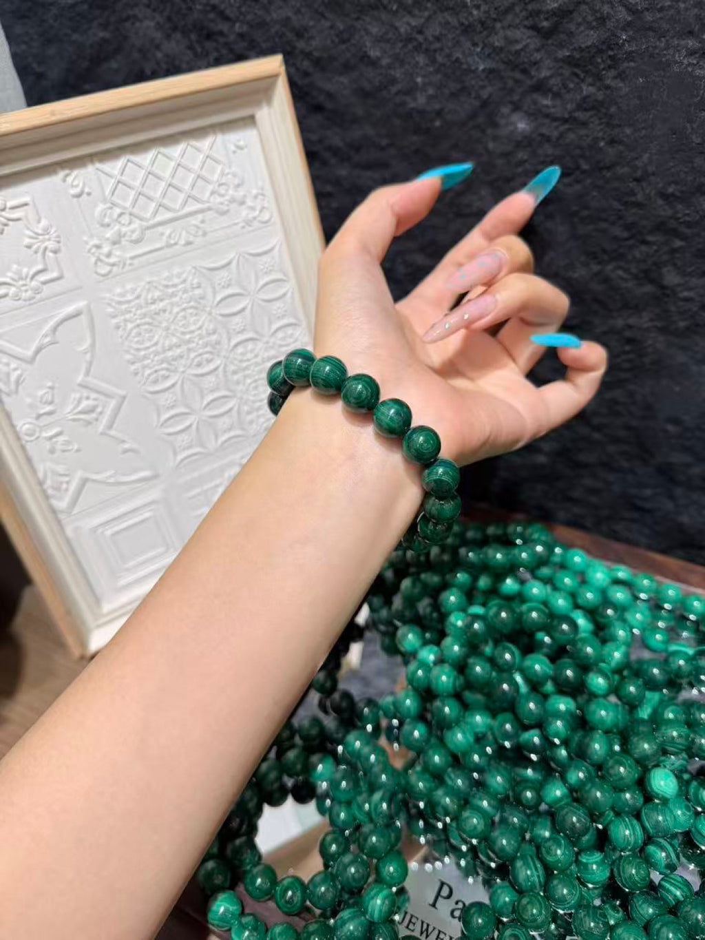 Wrist wearing a single-strand bracelet of round green malachite beads above a large pile of loose malachite beads, with a white decorative frame in the background