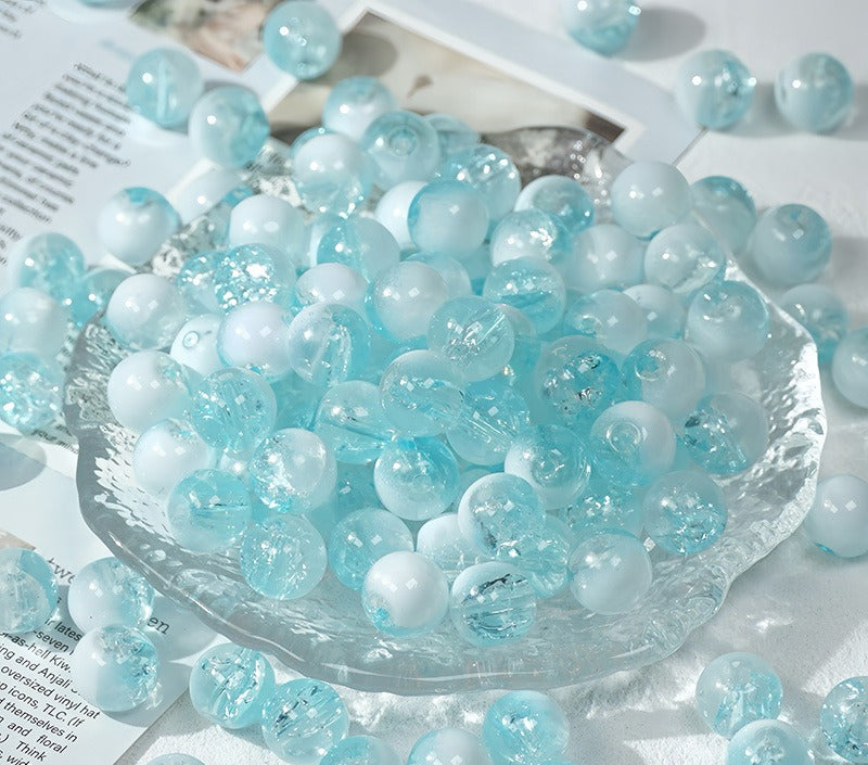 Light blue and white ice-crackle glass beads piled in a clear glass dish on a white surface