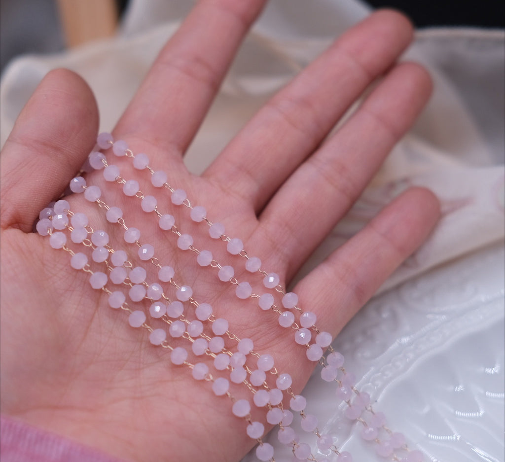 A close-up of a hand holding several strands of pale pink faceted beads with small gold spacers, draped over the palm against a soft beige fabric background.