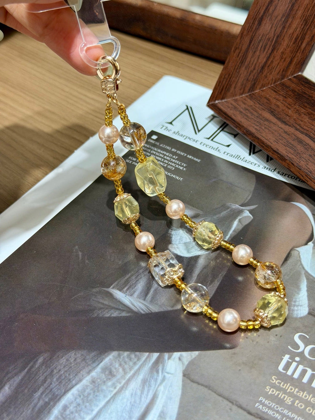 Hand holding a gold beaded phone charm with citrine crystals, pearls and clear glass cubes over a magazine page