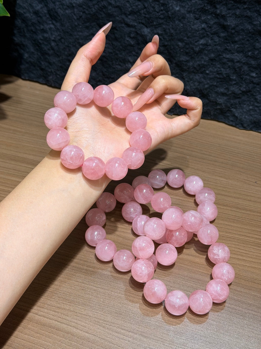 Wrist wearing a chunky pink quartz bead bracelet held above a wooden surface, with additional pink quartz bracelets coiled below