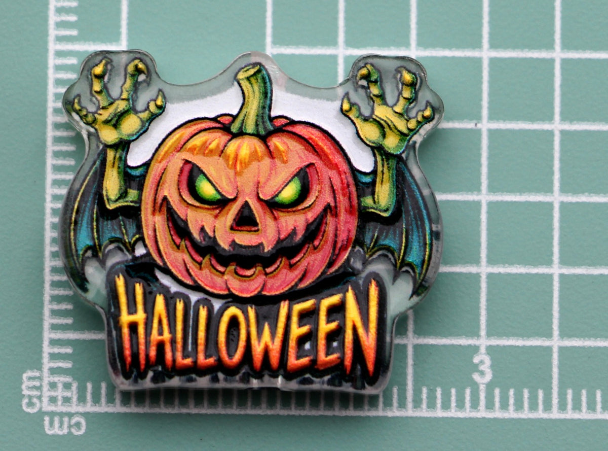 Halloween acrylic charm: carved orange pumpkin with green eyes, zombie hands and bat wings, above orange 'HALLOWEEN' text
