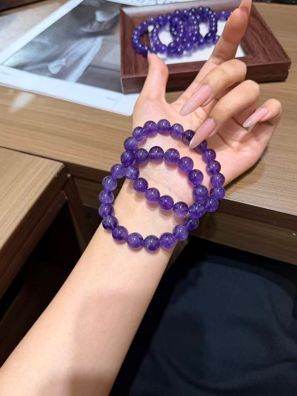 Hand holding a purple amethyst bead bracelet beside a wrist wearing a matching bracelet, with a wooden tray of bracelets and newspaper in the background