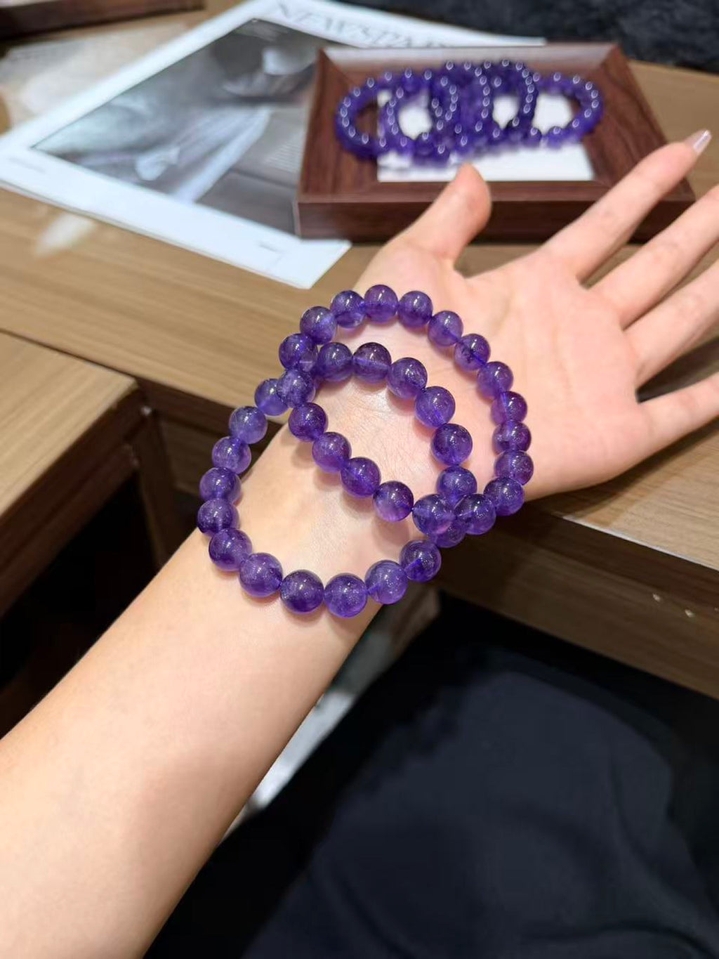 Two purple amethyst bead bracelets worn on a wrist, with more bracelets in a wooden display box in the background
