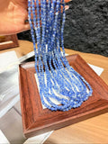 Hand holding long strands of small blue faceted kyanite beads draped over a wooden display tray.
