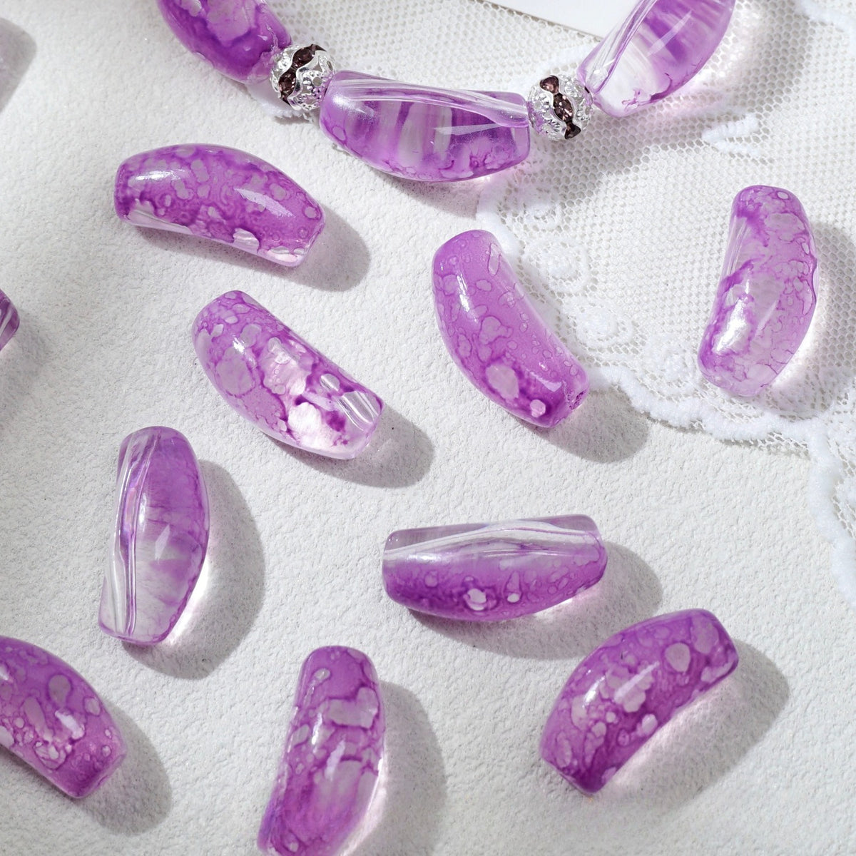 Purple marbled translucent curved glass tube beads scattered on white textured fabric with lace trim