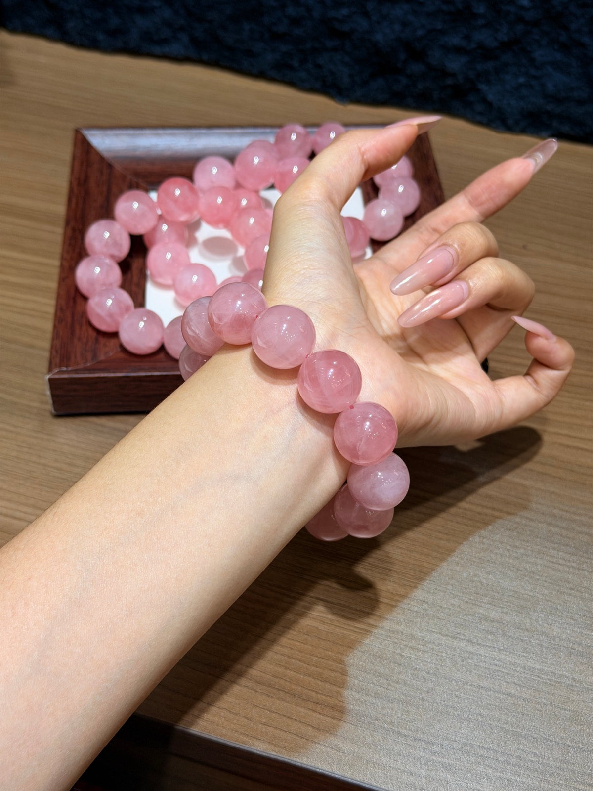 Wrist wearing a chunky pink quartz bead bracelet, with a wooden tray of pink quartz beads on a table behind