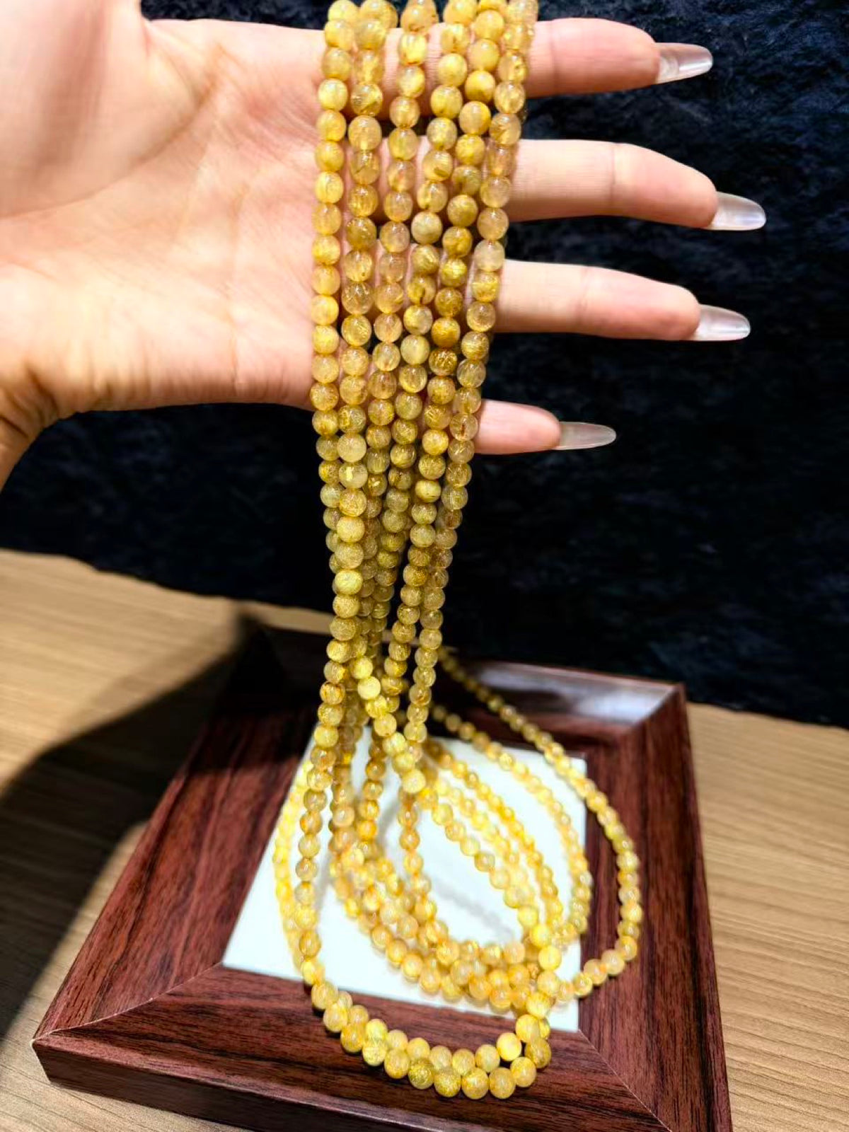 Hand holding long strands of golden rutilated quartz round beads draped over a wooden tray