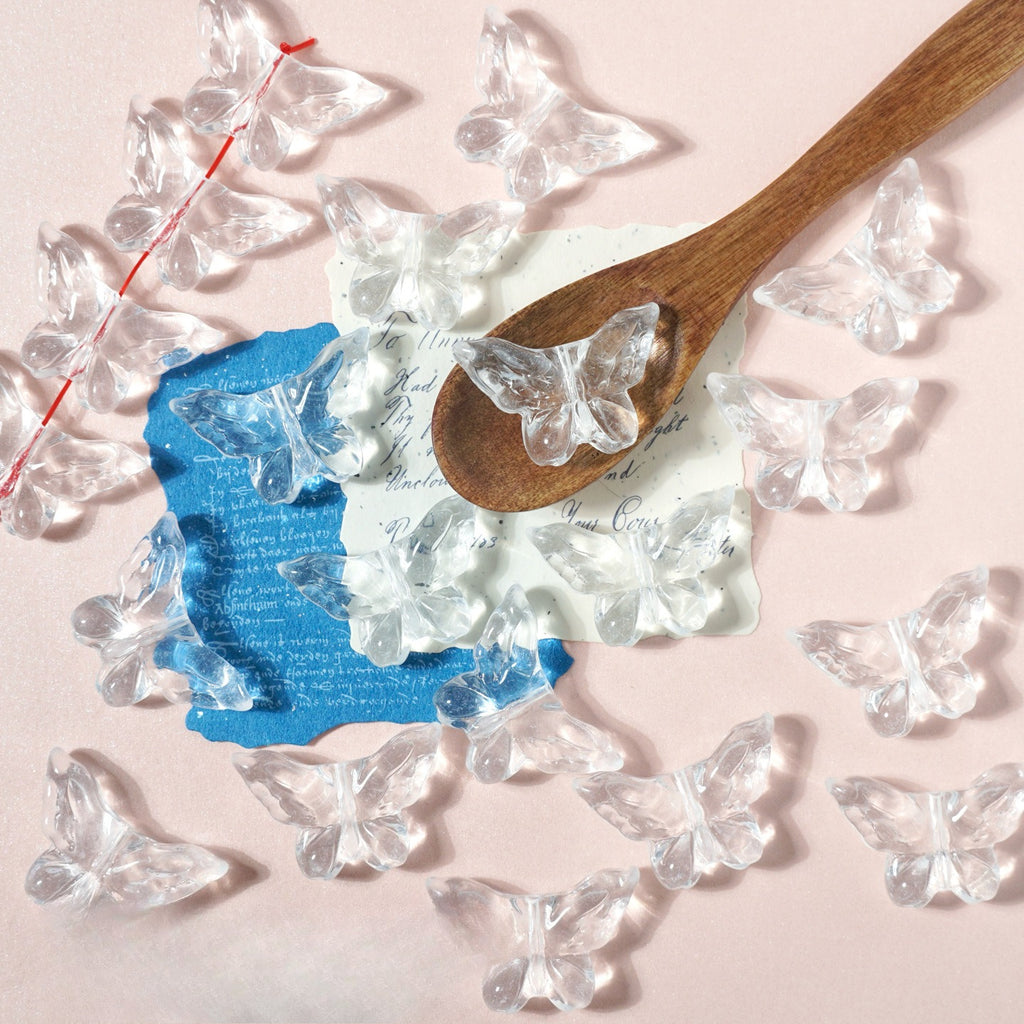 Clear butterfly-shaped glass charms on pale pink with a wooden spoon and blue paper accent