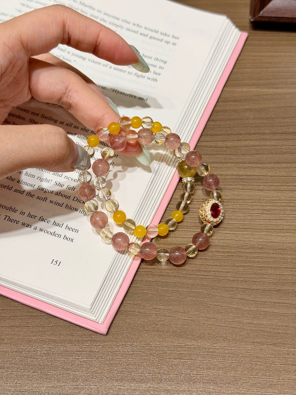 Hand holding a pink and yellow beaded bracelet with clear faceted beads and a gold charm with red stone over an open book