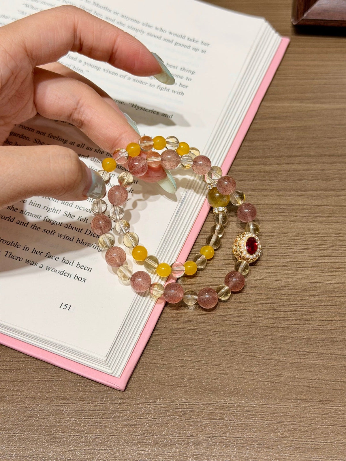 Hand holding a pink and yellow beaded bracelet with clear faceted beads and a gold charm with red stone over an open book