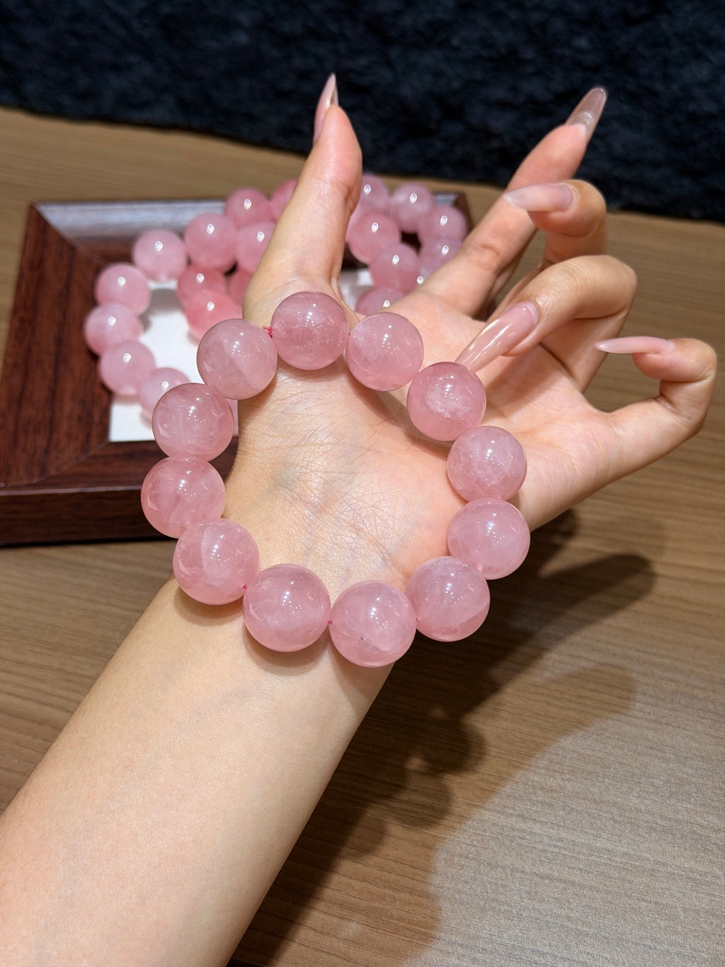 Chunky pink quartz bead bracelet draped across a hand, with a wooden tray of pink quartz beads on a table behind