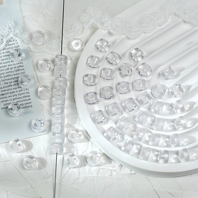 White glitter barrel beads on a white tray with lace fabric and a printed card behind