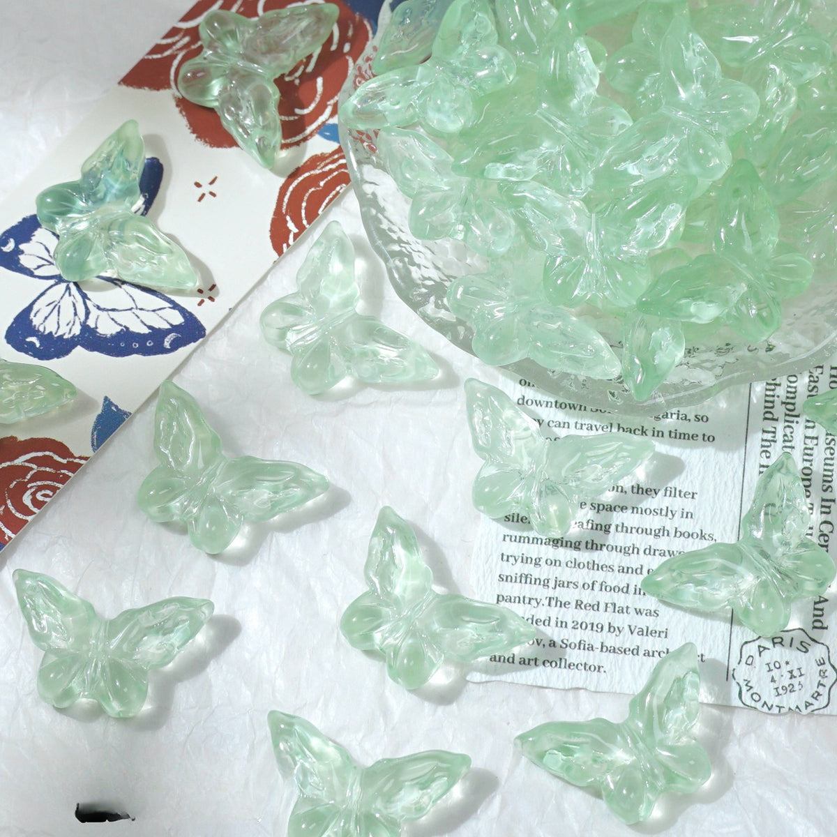 Light green translucent butterfly-shaped glass beads scattered on a white surface with a glass dish