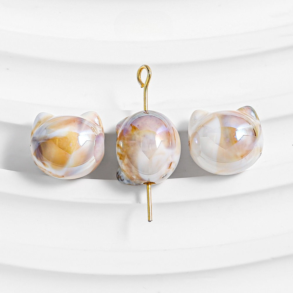 Three iridescent shell beads with cat-ear shapes and gold wire loops on a white curved surface.